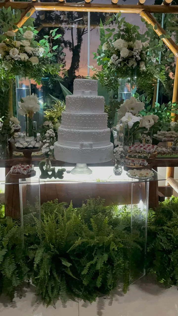 Mesa de bolo branco com flores brancas e samambaias sob o pergolado