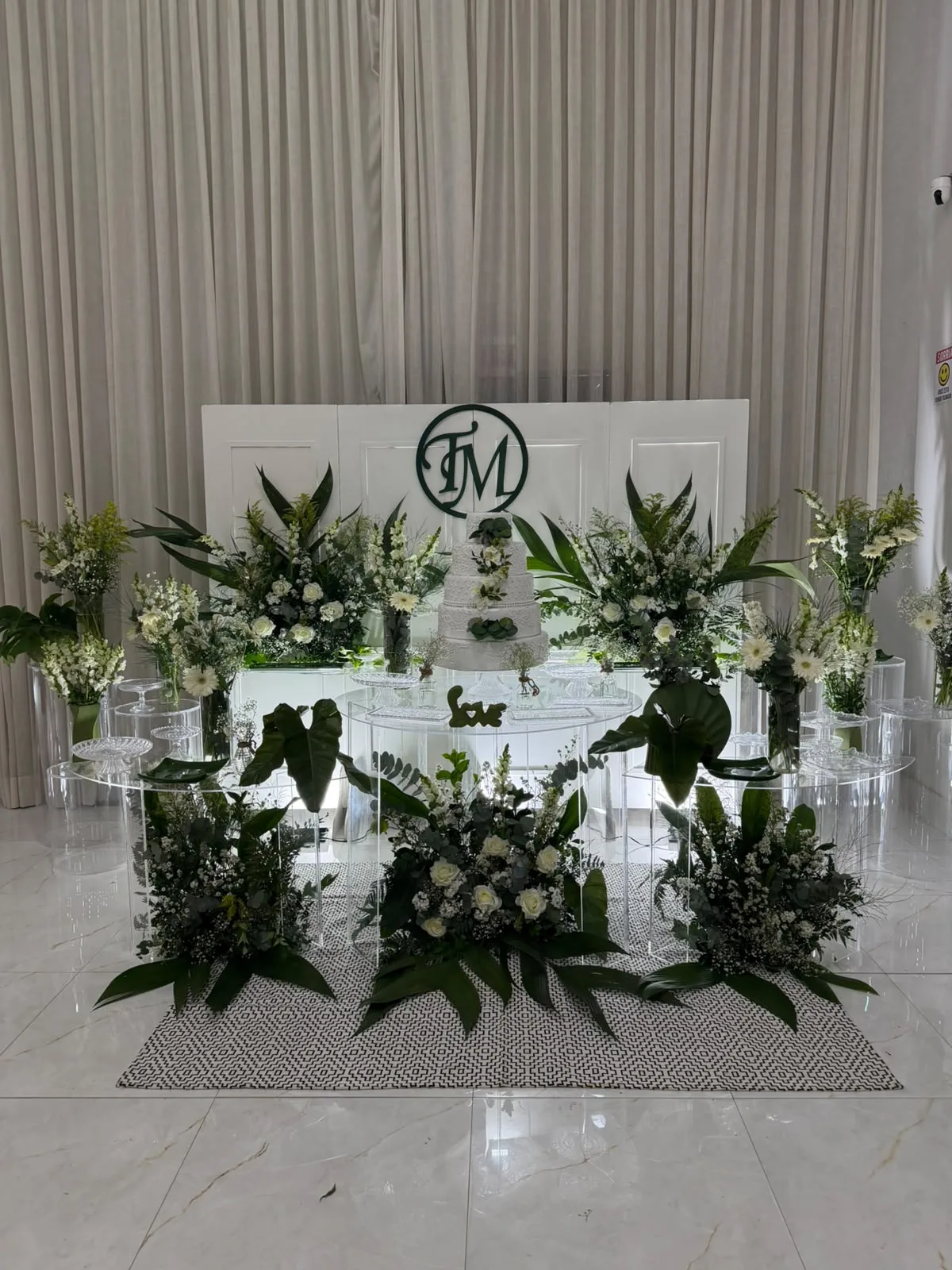 Mesa de bolo com folhagens e flores brancas diante de painel com monograma