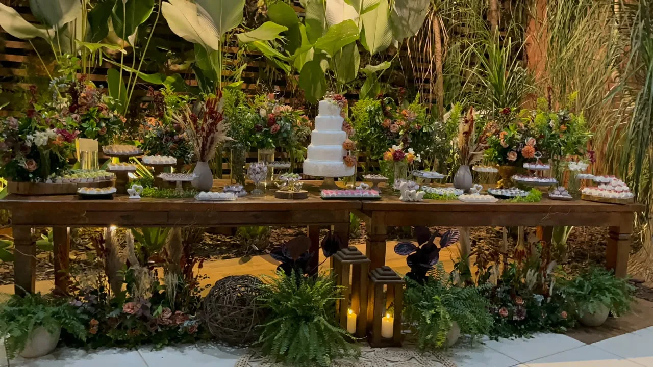 Mesa de doces rústica cercada de folhagens tropicais e lanternas